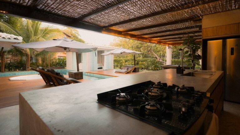 Área gourmet com churrasqueira e cooktop em bancada de pedra, pergolado e piscina ao fundo