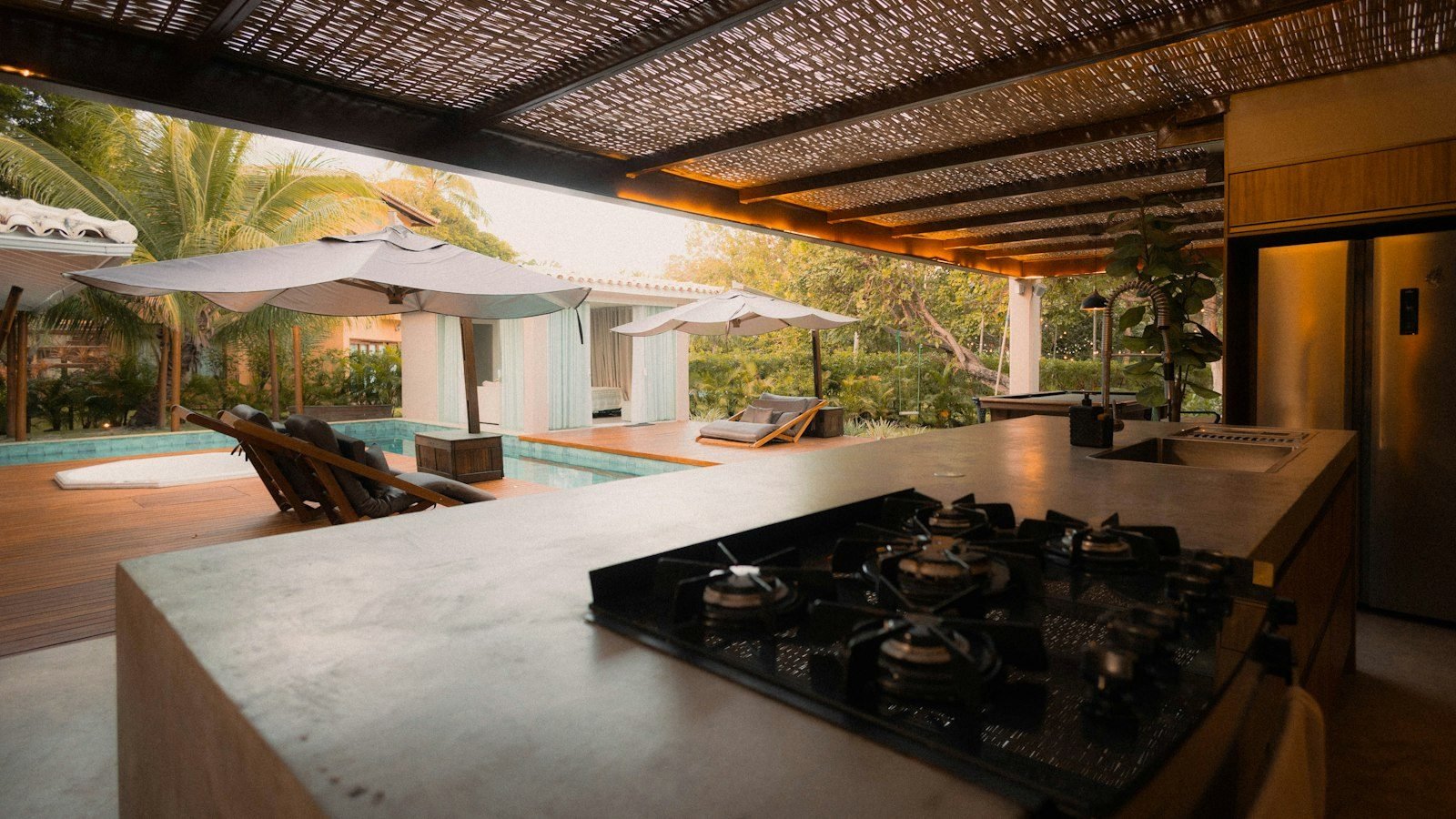 Área gourmet com churrasqueira e cooktop em bancada de pedra, pergolado e piscina ao fundo