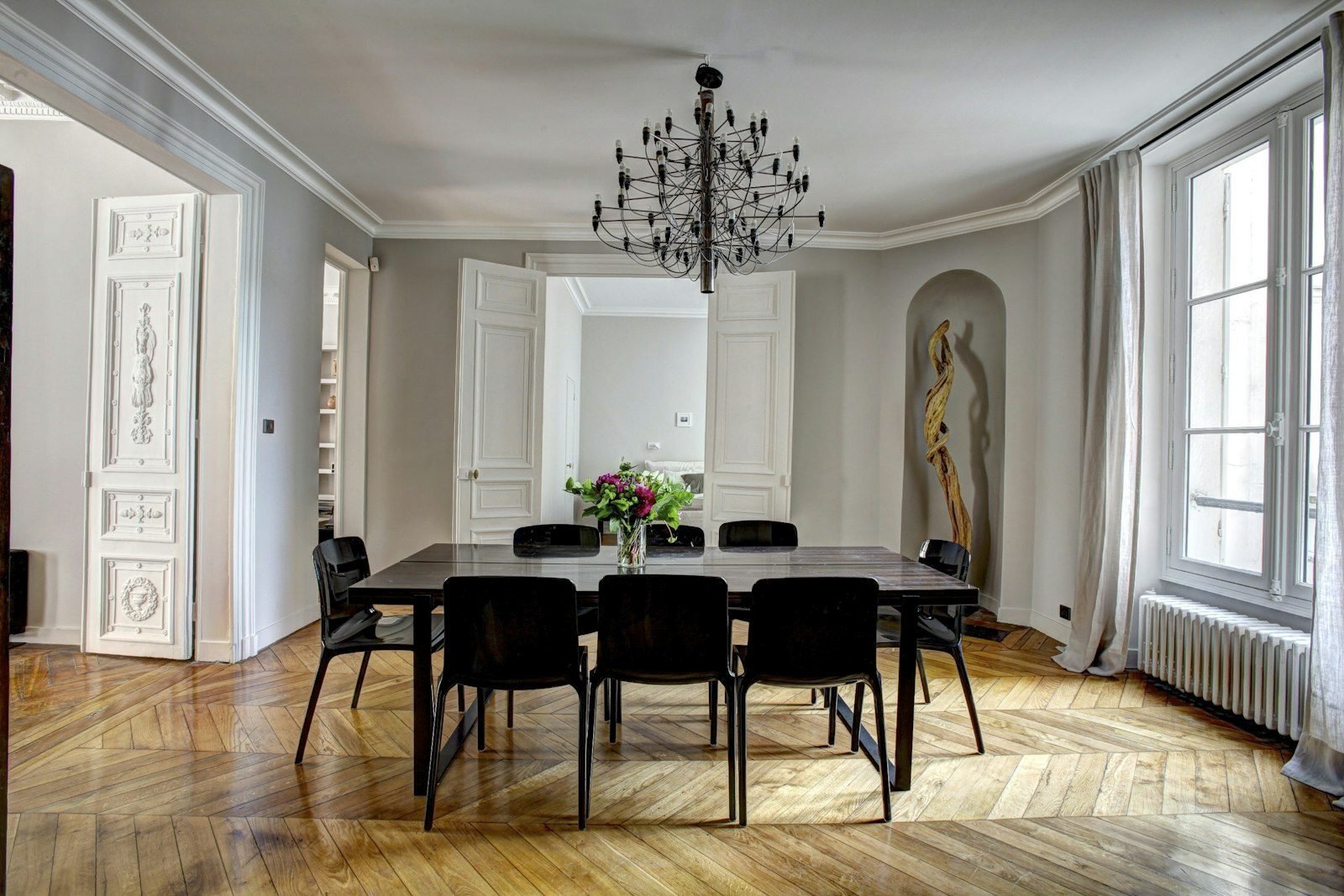 Sala de jantar clássica com mesa escura, cadeiras pretas, lustre de hastes, piso de madeira em espinha de peixe e janelas altas