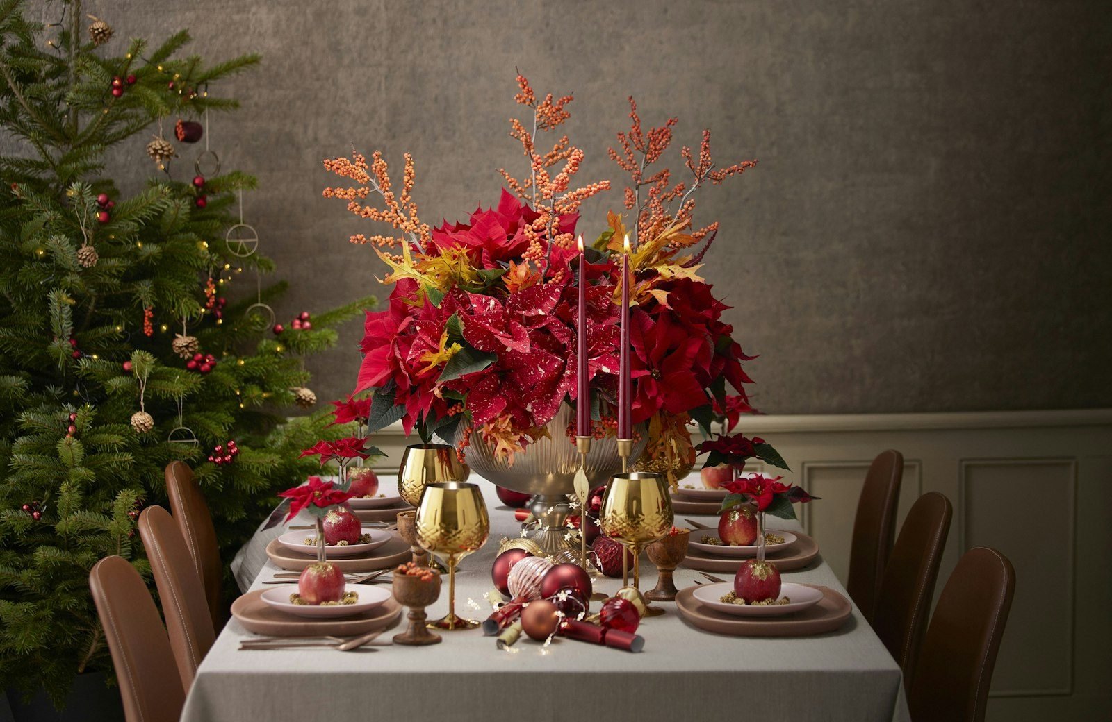 Mesa de Natal decorada com toalha branca, arranjo de bicos-de-papagaio vermelhos, taças douradas e árvore de Natal ao fundo