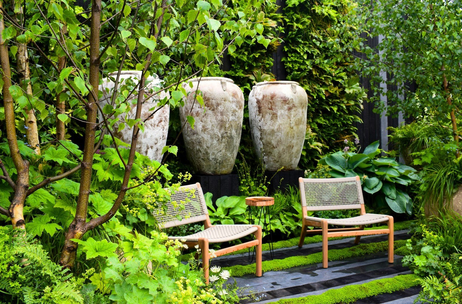 Jardim sombreado com duas poltronas de madeira entre folhagens e grandes vasos de cerâmica